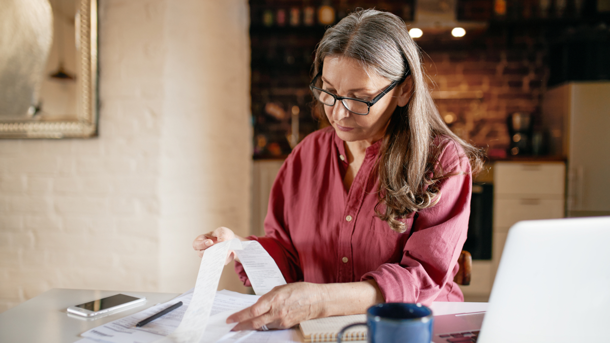 A woman wearing glasses reviews receipts and financial documents at a home office desk with a laptop, notebook, smartphone, and coffee mug, demonstrating organised bookkeeping and tax record management.